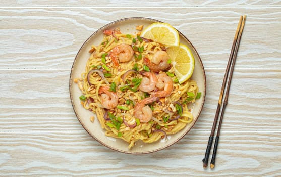 Pad thai noodles garnished with shrimp, peanuts, green onions, lemon wedges, and served with chopsticks on a white wooden table, Thai cuisine