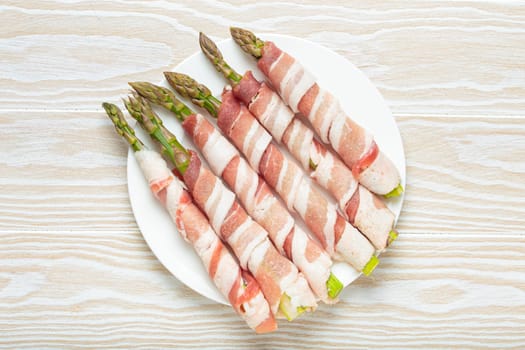 Delicious asparagus wrapped in bacon strips, seasoned with colorful peppercorns, presented on a white plate, ready to be cooked