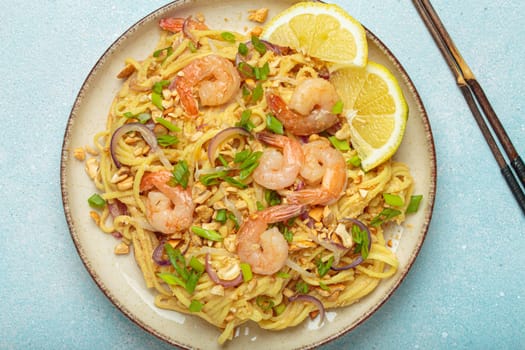 Pad thai noodles garnished with shrimp, peanuts, green onions, lemon wedges, and served with chopsticks on a light blue background, Thai cuisine