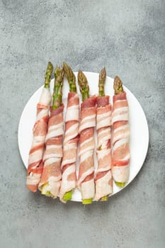 Delicious asparagus wrapped in bacon strips, seasoned with colorful peppercorns, presented on a white plate, ready to be cooked