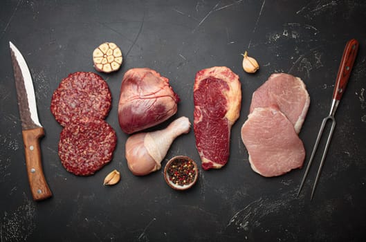 Selection of assorted raw meat cuts for zero carb carnivore diet: uncooked beef steak, pork, ground meat patty, heart, and chicken legs on black stone background top view with knife and carving fork