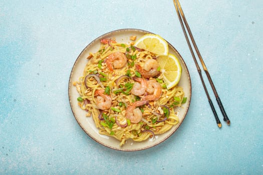 Pad thai noodles garnished with shrimp, peanuts, green onions, lemon wedges, and served with chopsticks on a light blue background, Thai cuisine