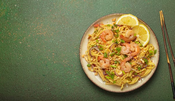 Pad thai noodles garnished with shrimp, peanuts, green onions, lemon wedges, and served with chopsticks on a rustic green background, Thai cuisine, copy space