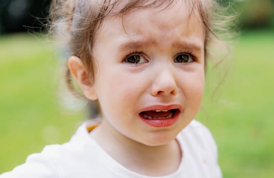 Portrait of one little beautiful Caucasian girl with brown eyes who looks away and cries bitterly on a spring day in the park, side view close-up with depth of field. Concept crying children.