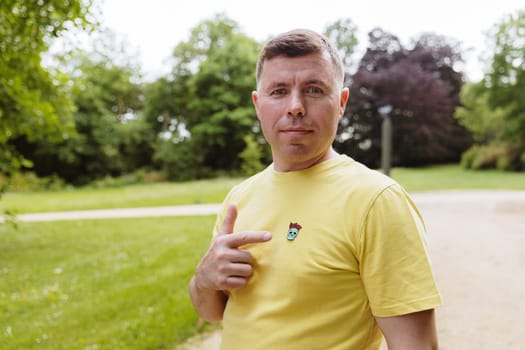 Portrait of one handsome Caucasian happy man in yellow T-shirt pointing finger at skull patch with roses standing in park on spring day, side view close up. Day of the Dead celebration concept.