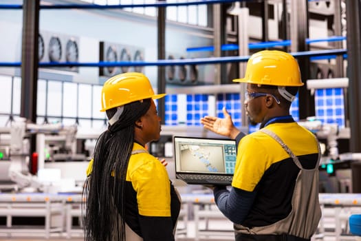 Black team of engineers supervising smart manufacturing process using digital interface on laptop. Robotic arms and automation assembly lines streamline industrial production for enhanced efficiency.