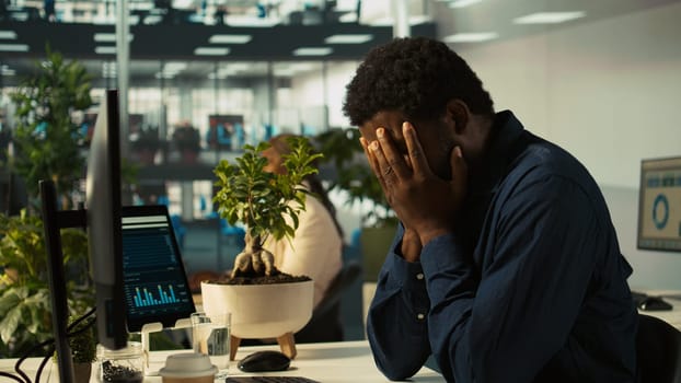 Overworked man having migraine symptoms while working on PC, drinking water to recover. Tired office worker in pain, feeling discomfort while doing tasks on PC, doing rehydration, camera B