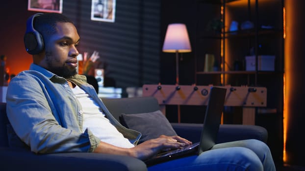 African american freelancer listening to a podcast being focused on online work, using headphones while seated on the couch in his living room. Multitasking in a casual home. Camera A.