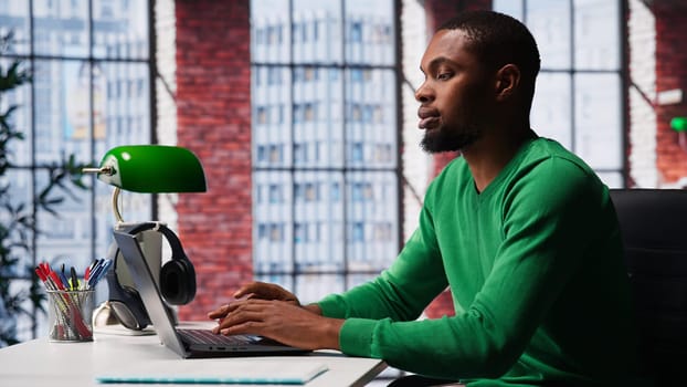 African american focused guy typing on his laptop at home office to solve daily tasks, doing remote freelance work online. Young adult surfing the web multitasking in loft apartment. Camera A.