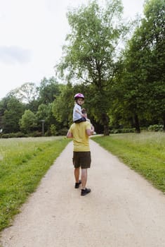 Vertical portrait from the back of one handsome Caucasian young man who holds on his shoulders a little girl with a bicycle helmet on her head, walking along a path in the park on a spring day, side view, distant plan. Concept happy childhood, time family walk park.