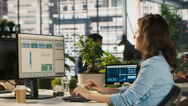 Accountant in busy modern startup agency imputing financial figures on PC after checking reports. Focused businesswoman working in modern office, crosschecking data, camera A
