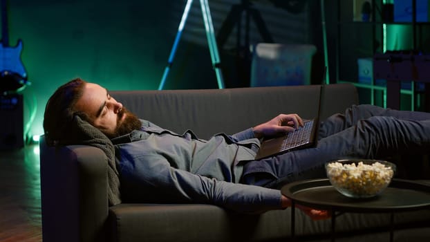 Exhausted man sleeping on couch after working on laptop all day before managing to watch TV and eat popcorn. Freelancer falls asleep while solving tasks on notebook and watching film, camera A