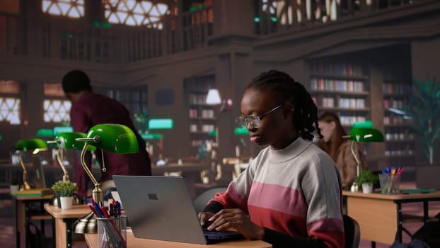 African american student consulting academic research collection at library, studying for upcoming exams to obtain her university diploma. Girl works on a scholarship, education. Camera B.