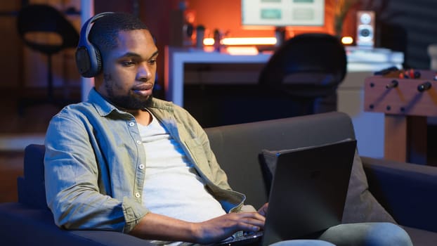 African american freelancer listening to a podcast being focused on online work, using headphones while seated on the couch in his living room. Multitasking in a casual home. Camera B.
