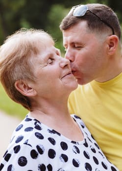 Portrait of one young Caucasian man lovingly kissing an elderly woman on the cheek while standing in the park on a spring sunset day, side view medium shot. Concept kiss day, mother's day, happy people.