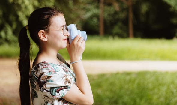 Portrait of a beautiful Caucasian girl with glasses standing sideways taking a picture of a landscape with a camera in a park on a spring sunset day, side view close-up with space for text on the right. Happy childhood concept, children bloggers.