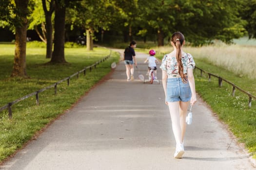 Portrait of three little Caucasian girls: one is walking with a camera in her hand, the second is riding a bicycle, and the third is walking with badminton rackets along a path in the park on a spring sunset day, side view, general plan with selective focus. Happy childhood concept, children are riding bicycles.