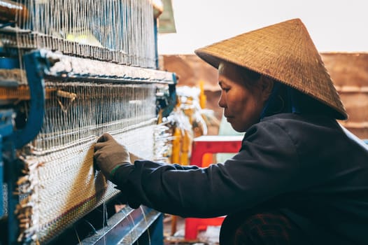 Vietnamese woman weaver in straw hat works at a loom weaving machines in workshop at factory for production of woven reed mats in Asia. An Cu, Tuy An, Phu Yen province, Vietnam - April 14, 2025