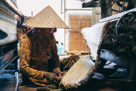 Vietnamese woman weaver in straw hat works at loom weaving machines in workshop at factory for production of woven reed mats in Vietnam. An Cu, Tuy An, Phu Yen province, Vietnam - April 14, 2025