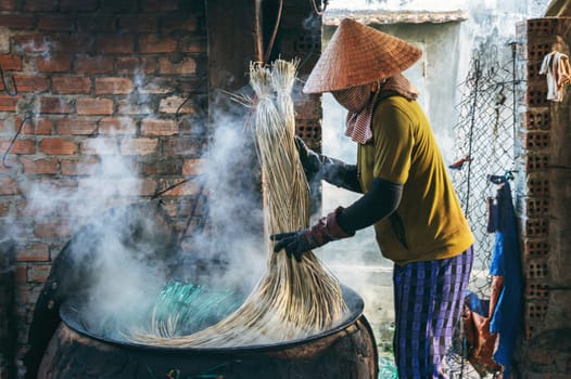 Vietnamese woman in a traditional straw hat dyes reeds at woven mat factory in Vietnam