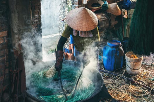 Vietnamese woman in a traditional straw hat dyes reeds at woven sleeping mat factory in Vietnam
