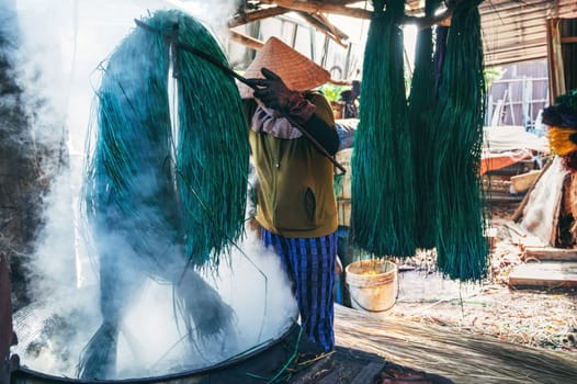 Vietnamese woman in a traditional straw hat dyes reeds at woven mat factory in Vietnam