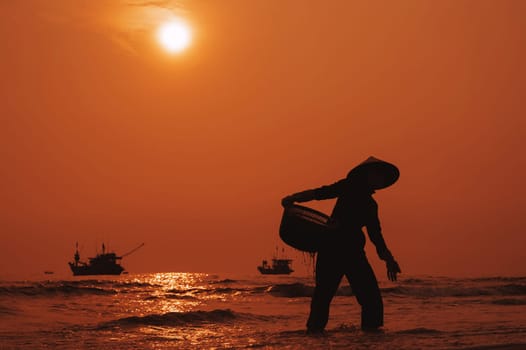 Silhouette of Vietnamese woman in straw hat on beach of sea with ships in fishing market in village in the morning at sunrise in Vietnam
