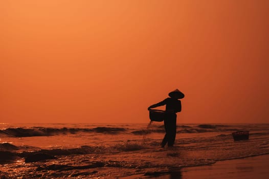 Silhouette of a Vietnamese woman in a straw hat on beach of sea in a fishing village in the morning at sunrise in Vietnam