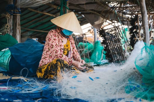 Vietnamese woman in traditional straw hat Non La repairs fishing net in a workshop on beach in fishing village in Vietnam