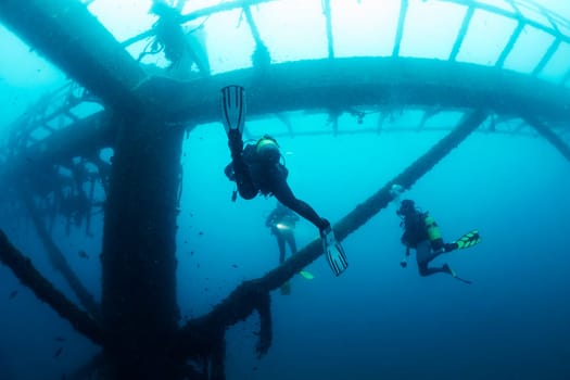 unrecognizable divers diving next to the structure of a sunken wreck in the sea, concept of scuba diving and underwater adventure