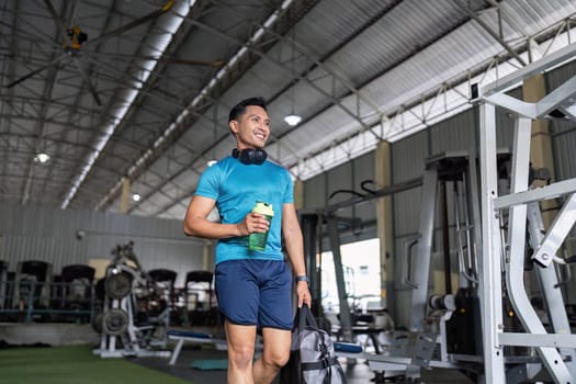A smiling man carrying a water bottle and gym bag, walking through a spacious gym. Emphasis on fitness lifestyle and positivity.