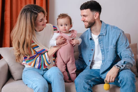 Young parents holding their baby while sitting on a sofa, smiling and enjoying a cozy moment together. Warm indoor family scene filled with joy, love, and parental connection.