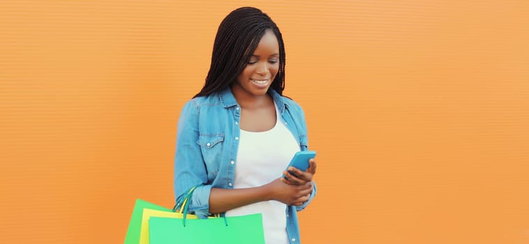 Beautiful happy African young woman holding phone with shopping bag, stylish black girl using smartphone wearing casual clothes on orange background