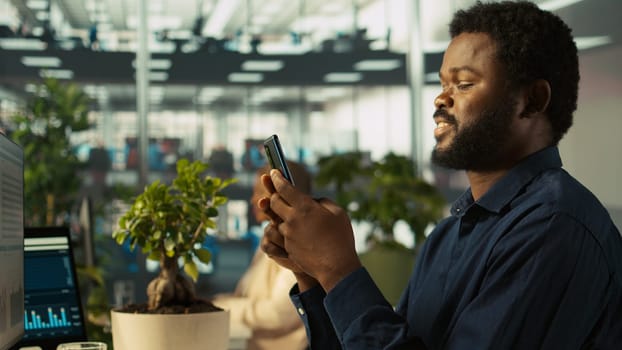 Employee in office taking break from checking documents to text friends using smartphone, relaxing. Accountant in office pausing tasks to answer messages on mobile phone, camera B