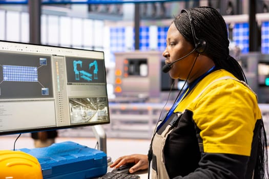 African american technician monitors automated factory conveyor belt and robotic arms, ensuring precision in industrial plant. Professional engineer uses computer in manufacturing solar panels.