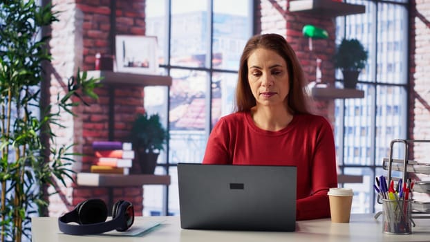 Mature girl freelancer managing her creative projects remotely in loft, working on a new novel or script for her gig. Woman focusing on writing and editing her creation, telework. Camera A.