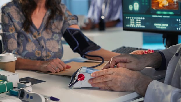 Senior physician measuring blood pressure with a blood pressure monitor, using professional tools at clinical consultation. Old doctor does a tension reading and heartbeat measurement. Camera B.