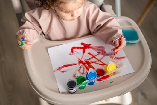 Top view of cute toddler girl painting with gouache at a table near a sunlit window