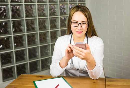 Happy Female Medical Doctor Sitting at her Table and Browsing Something on her Mobile Phone.