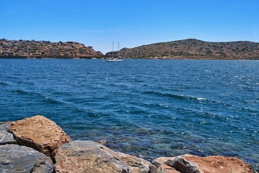 Spinalonga island, Venetian fortress and anchored yacht in lagoon with breakwater stones in foreground on clear sunny summer day, Crete, Greece. film grain