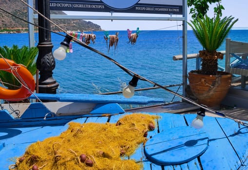Outdoor installation of traditional fisherman's boat, fishing net and octopuses clipped to rope to dry in sun in selective focus and calm blue sea and typical Greek hills in background, Crete, Greece. Translation German, French, Russian to English: grilled octopus