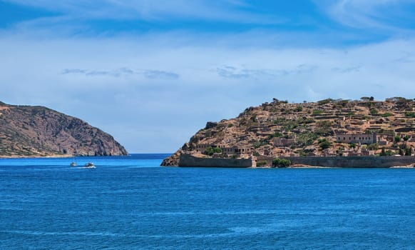 Beautiful view of west bank of Spinalonga island and Venetian fortress on clear sunny summer day on Crete, Greece. Boats passing, old fortifications, calm blue sea.