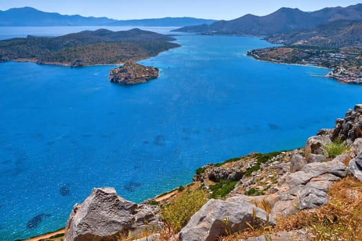 Aerial view of clear sea waters of harbor between Spinalonga island and peninsula, known as Kalydon, and Plaka village from above and rocky slope in foreground, Crete, Greece. film grain