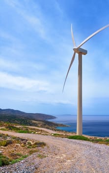 Single windmill turbine on hilltop of seashore in colorful landscape against dynamic blue sky with clouds and winding road. Vertical shot. Crete, Greece.
