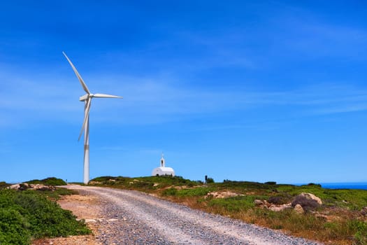 Single windmill turbine and small traditional Greek Orthodox chapel or church and road to them in colorful landscape on hilltop against blue sky with some cloulds on clear sunny summer day. Crete, Greece