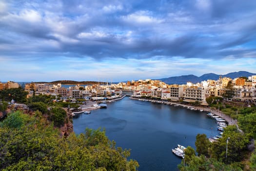 Colorful view of Voulismeni lake and Agios Nikolaos town on Crete island, Greece in cloudy spring evening. Boats, cafes, restaurants, ship, marina, promenade.