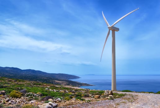 Single windmill turbine on hilltop of seashore in colorful landscape against dynamic blue sky with clouds and winding road. Crete, Greece.