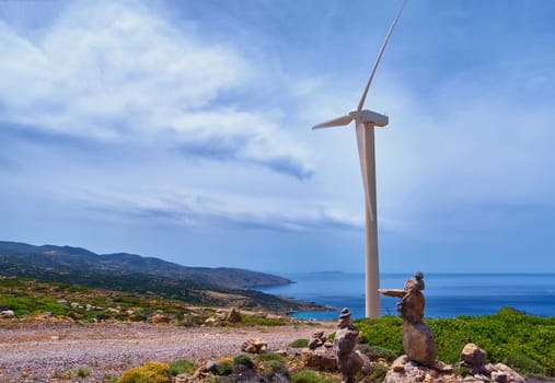 Single windmill turbine and balancing stones on hilltop of seashore in colorful landscape against dynamic blue sky with clouds and winding road on clear sunny summer day. Crete, Greece.