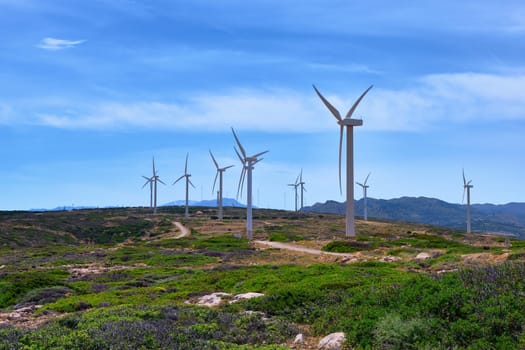 Windmill turbine farm on a hilltop in colorful landscape with winding road against blue sky on clear sunny summer day. Crete, Greece.