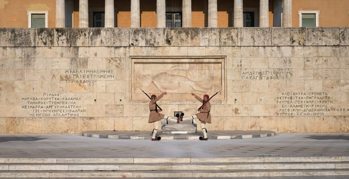Ceremony of Presidential guard Evzones by the Greek tomb of Unknown Soldier. Evzones in summer uniform face each other, hands up, legs up. Parliament building. Wide shot.